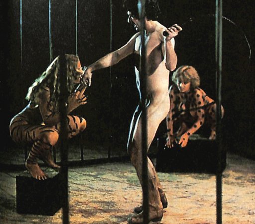 naked man with a whip training animal-painted women locked in a circus cage